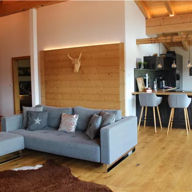 A modern living room with a gray sofa and wooden details. In the background, an open kitchen with bar stools is visible.