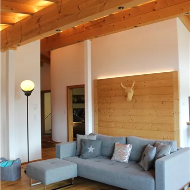 A modern furnished living room with a gray sofa and a wooden floor. The ceiling is designed with wooden beams, and there is cozy lighting.