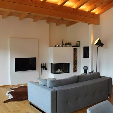 A modern living room with a gray sofa and a fireplace. The room has wooden beams and large windows that provide plenty of light.