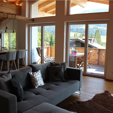A modern living room with a gray couch and large windows. In the background, an open kitchen and a balcony with a view of the mountains are visible.