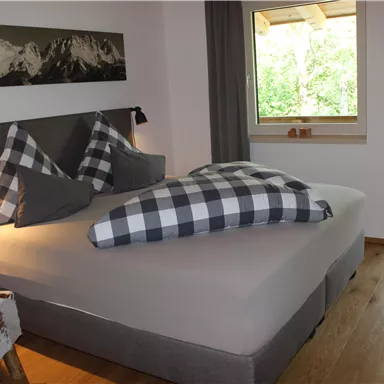 A modern bedroom with a large bed and checked pillows. Large windows offer a view of greenery and plenty of daylight.