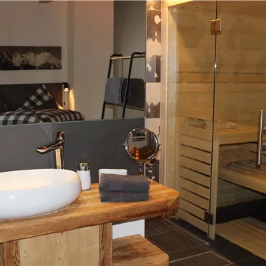 A modern bathroom with an elegant ceramic sink and a wooden table. Next door is a wooden sauna with a glass door.