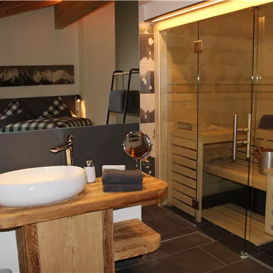 A modern bathroom with a stylish sink and a large mirror. In the background, there is a wooden sauna and a glass shower cabin.