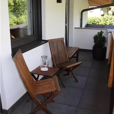 A cozy balcony with two wooden chairs and a small table. In the background, you can see green trees and plenty of daylight.