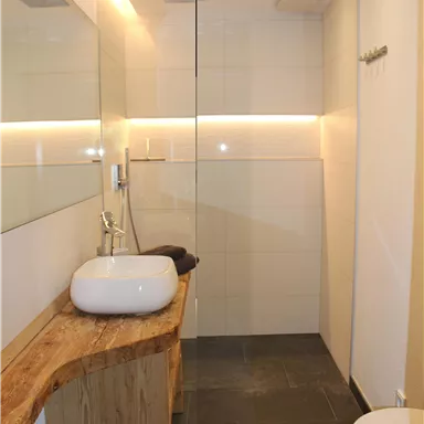 A modern bathroom with a glass door shower and elegant white walls. The wash area features a wooden washbasin and a stylish sink.