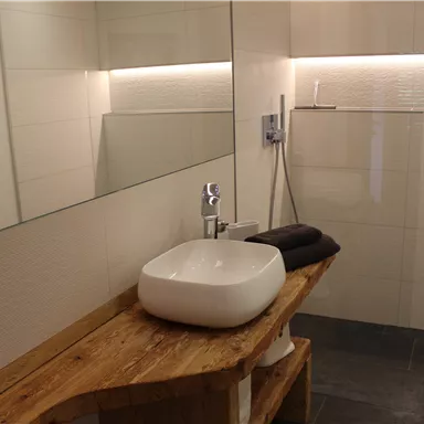 A modern bathroom with a white sink and a wooden shelf. The walls are light and there is a large mirror surface.
