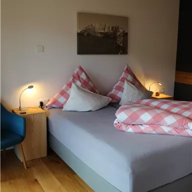 A cozy bedroom with a large bed and checkered pillows. Next to the bed is a small wooden table with a lamp and a blue armchair.