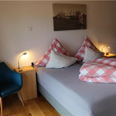 A cozy bedroom with a large bed and checked pillows. There is a small table with a lamp and a blue armchair next to it.