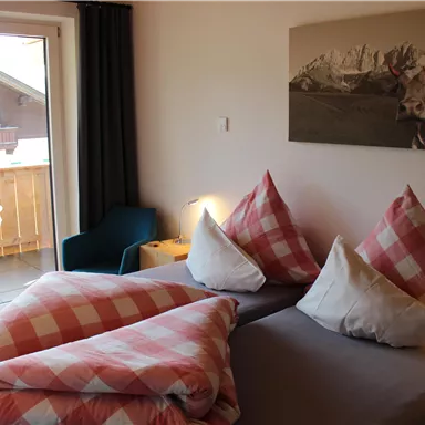A cozy bedroom with two beds and checked pillows. The room has a balcony with a view of the mountains.