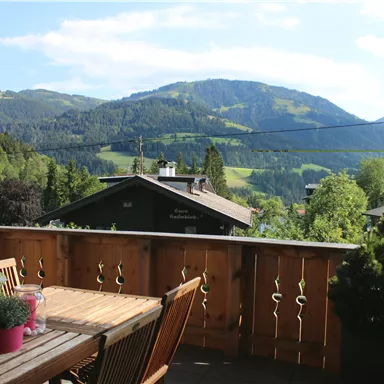 A beautiful balcony with wooden furniture and flowering plants. In the background, green mountains and a clear sky can be seen.