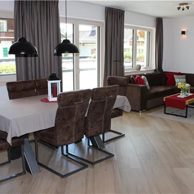 Eine moderne und einladende Wohnküche mit einem Esstisch und bequemen Sitzgelegenheiten. Große Fenster lassen viel Tageslicht herein und schaffen eine helle Atmosphäre.