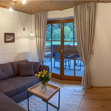 A cozy living room with a large gray couch and a wooden table. Large windows offer a view of a balcony and the green surroundings.