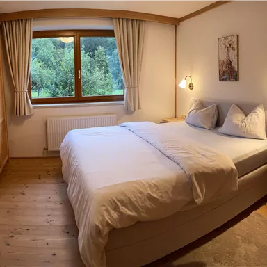 A cozy bedroom with a large bed, soft colors, and wooden details. Through the window, a view of greenery is visible.