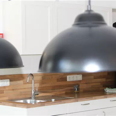 Eine moderne Küche mit Holzdetails und eleganten, schwarzen Pendelleuchten. Im Hintergrund sind Geschirr und Geräte sichtbar.