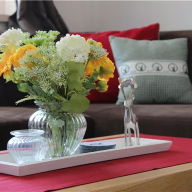 Ein stilvoll dekorierter Tisch mit einer Blumenvase und einem kleinen Dekorationsobjekt. Im Hintergrund sind ein gemütliches Sofa und Kissen zu sehen.