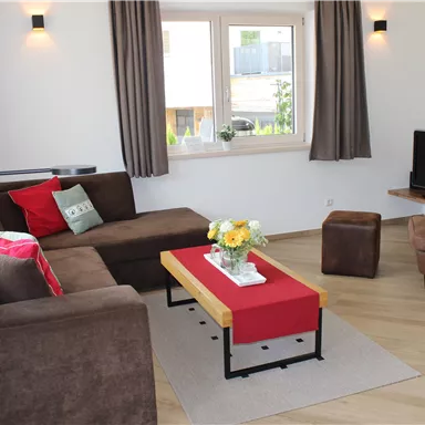 Ein gemütliches Wohnzimmer mit einem braunen Sofa und einem roten Tisch. Es gibt einen Fernseher und viel Tageslicht durch die Fenster.