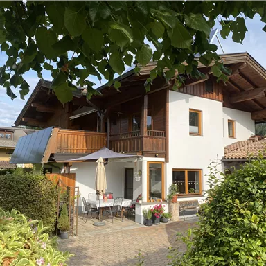 A cozy house with a large balcony and a garden. In the background, trees and mountains are visible.