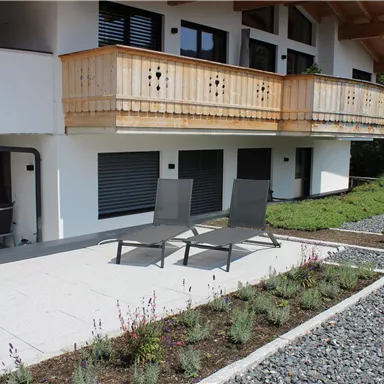Ein modernes Haus mit einem Balkon und einer Terrasse. Auf der Terrasse stehen Liegestühle und umgeben sind sie von schönem Garten.