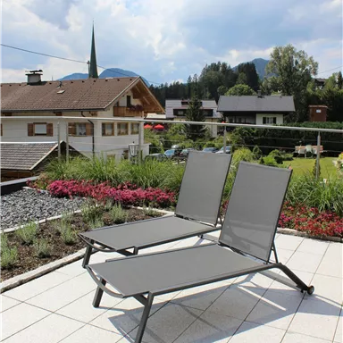 Zwei graue Liegen auf einer Terrasse mit blauen Himmel und Wolken. Im Hintergrund sind Häuser und Bäume sichtbar.
