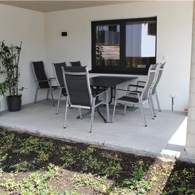 Ein einladender Patio mit einem Tisch und sechs Stühlen. Im Hintergrund stehen einige Pflanzen und es gibt viel Tageslicht.