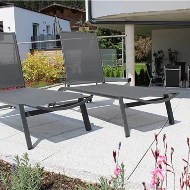 Zwei moderne Liegestühle auf einer Terrasse. Im Hintergrund sind Pflanzen und ein gemütlicher Sitzbereich sichtbar.