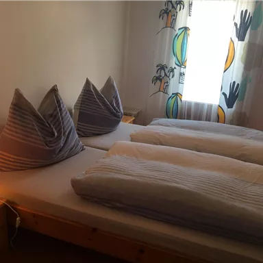 A cozy bedroom with two single beds and decorative pillows. The room has a warm tone and a light curtain with a colorful pattern.