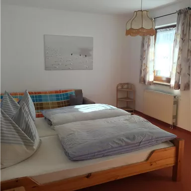 A simple bedroom with a double bed and two large pillows. The room has bright walls and a large window with curtains.
