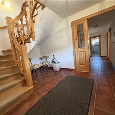 Ein einladender Flur mit einer Holztreppe und Fliesenboden. Auf der linken Seite befinden sich einige Stühle und eine Tür, die zu weiteren Räumen führt.