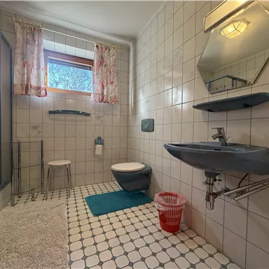 Ein Badezimmer mit gefliesten Wänden und einem großen Fenster. Es gibt eine Dusche, ein Waschbecken und eine Toilette, sowie einige Badezimmeraccessoires.