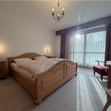 Ein gemütliches Schlafzimmer mit einem Doppelbett aus Holz und einer sonnigen Fensterfront. Der Raum ist hell und lädt zum Entspannen ein.