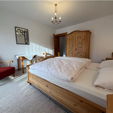 Ein gemütliches Schlafzimmer mit einem großen Bett und einem Holzschrank. Ein roter Sessel und ein kleiner Tisch runden die Einrichtung ab.