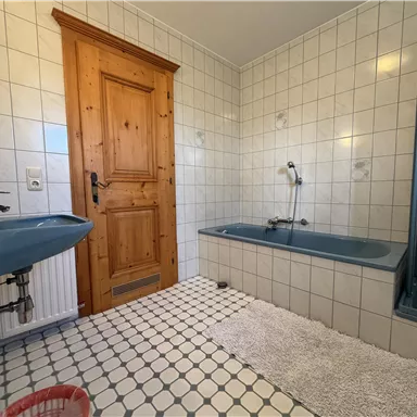 Ein Badezimmer mit weißen Fliesen und blauen Sanitäranlagen. Es gibt eine Badewanne, eine Dusche und eine Holz-Tür.