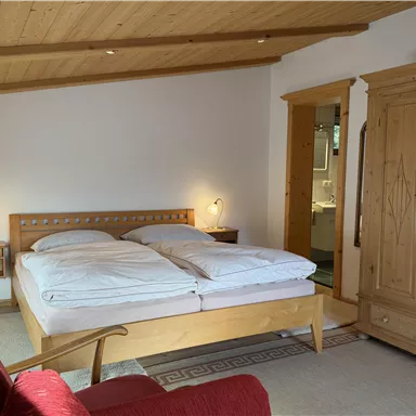Ein gemütliches Schlafzimmer mit einem großen Bett, zwei Nachttischen und einem roten Sofa. Die Wände sind hell, und es gibt einen rustikalen Kleiderschrank aus Holz.