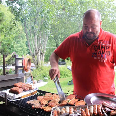 A man is grilling on a charcoal grill with various types of meat. In the background, green trees and a natural environment can be seen.