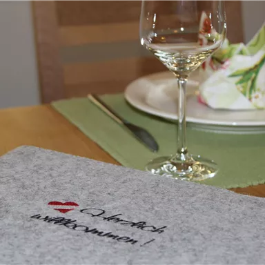Ein schöner Tisch gedeckt mit einem Glas Wein und einem dekorierten Teller. Auf dem Tisch liegt eine graue Stoffmappe mit dem Schriftzug „Willkommen!“.
