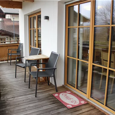 Ein gemütlicher Balkon mit Holzdielen, zwei Stühlen und einem Tisch. Durch die großen Fenster ist ein schöner Blick nach draußen möglich.