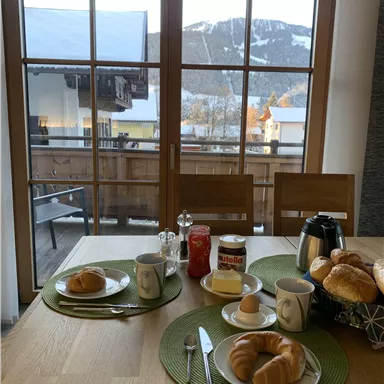 Ein gemütlicher Frühstückstisch mit Brötchen, Marmelade und Getränken. Im Hintergrund sind schneebedeckte Berge und ein Balkon zu sehen.