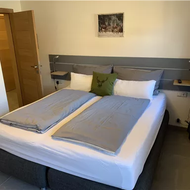 Ein gemütliches Schlafzimmer mit einem großen Doppelbett und modernen Möbeln. An der Wand hängt ein Bild und es gibt Fenster mit Vorhängen.