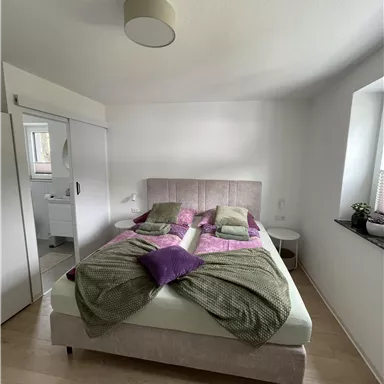 A modern bedroom with a large double bed and colorful pillows. The room is bright and cheerful, with a window that lets in a lot of light.