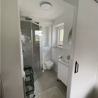 A modern bathroom with a shower, toilet, and sink. The walls are light and there is a large window for natural light.