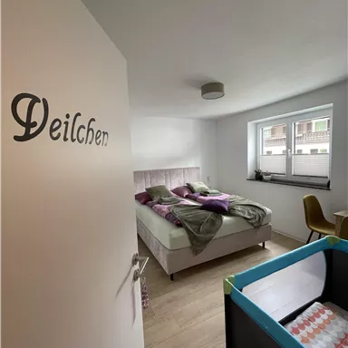A modern bedroom with a double bed and a baby curve. There are large windows that provide plenty of daylight.
