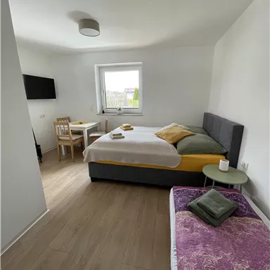 A bright room with a large bed and a cozy seating area.
There is a table and a bench as well as a television on the wall.