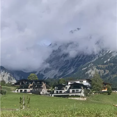 Eine malerische Landschaft mit modernen Häusern in einer grünen Wiese. Im Hintergrund ragen beeindruckende Berge und bewölkter Himmel hervor.