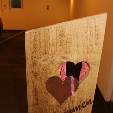 Ein Holzschild mit der Aufschrift "Willkommen" und Herz-Ausschnitten. Im Hintergrund ist ein gut beleuchteter Parkplatz zu sehen.