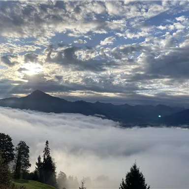 Eine atemberaubende Aussicht auf Berge, die von Nebel umhüllt sind. Der Himmel ist mit Wolken und Sonnenstrahlen gefüllt.