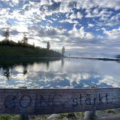 Eine Holzbank am Ufer eines ruhigen Gewässers mit der Aufschrift "GOING stärkt". Im Hintergrund spiegeln sich Wolken im Wasser und die Landschaft ist sanft beleuchtet.