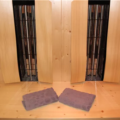 Ein entspannender Raum mit zwei Infrarotstrahlern und Holzverkleidung. Zwei violette Handtücher liegen auf dem Boden.