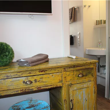 Ein gemütlicher Raum mit einem gelben, vintage Möbelstück und einem Fernseher an der Wand. Im Hintergrund ist ein kleines Badezimmer mit Waschbecken zu sehen.