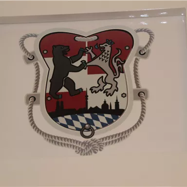 Ein Wappen mit einem Bären und einem Löwen, umrahmt von rot-weißem Design und blauen Rauten. Es stellt emblematische Symbole dar, die mit Städten oder Regionen verbunden sind.