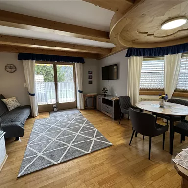 A bright and inviting kitchen with wooden ceilings and a cozy atmosphere. The room features a dining table, a television, and large windows for plenty of natural light.